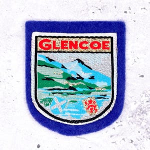 May include: A blue felt patch with an embroidered design. The design features the word "GLENCOE" in red letters above a landscape scene with mountains, water, and a Scottish flag. The patch has a white border and a blue felt backing.