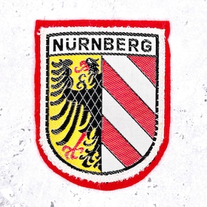 Peut inclure: Un écusson brodé rectangulaire à bordure rouge portant le texte NURNBERG en haut. Le blason est divisé verticalement, affichant un aigle noir sur fond jaune à gauche et des bandes diagonales rouges et blanches sur la partie droite.