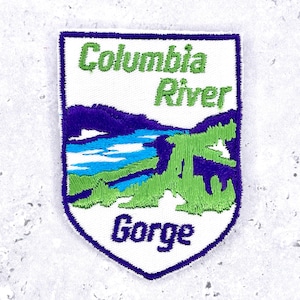 May include: A shield-shaped embroidered patch featuring a white background with a landscape of blue water, green hills, and dark purple mountains. The text Columbia River is stitched in green, and the word Gorge is stitched in dark purple.