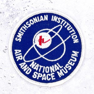 May include: A circular embroidered patch with a dark blue background and a white border. It features a white planet graphic with a red satellite icon and white orbital rings. The text reads Smithsonian Institution National Air and Space Museum.