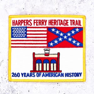 May include: A square embroidered patch with a yellow border. It features the text HARPERS FERRY HERITAGE TRAIL at the top and 260 YEARS OF AMERICAN HISTORY at the bottom. The centre displays a US flag, a Confederate flag, and a red brick building.