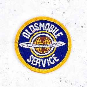 May include: Round patch with a blue background and yellow border. The patch features the words "OLDSMOBILE SERVICE" in white, surrounding a brown globe with a white ring. A vintage automotive emblem.