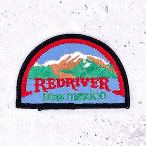 Może przedstawiać: Haftowana naszywka z motywem górskiego krajobrazu. Naszywka przedstawia niebieskie niebo, zielone i brązowe góry oraz napisy "RED RIVER new mexico" w kolorze czerwonym i zielonym. Naszywka ma czarną obwódkę.
