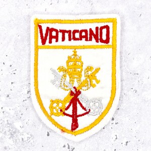 May include: A shield-shaped embroidered patch featuring the word VATICANO in red capital letters at the top. Below the text, the design displays a yellow papal tiara above two crossed keys, one in yellow and one in silver, tied with red cords.