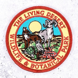 May include: A circular embroidered patch with a red border and cream background featuring the text THE LIVING DESERT WILDLIFE & BOTANICAL PARK. The center depicts a coyote, a bighorn sheep, a tortoise, an owl, and a snake under a yellow sun.