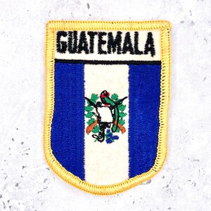 May include: A rectangular embroidered patch featuring the flag of Guatemala with vertical blue and white stripes. The word GUATEMALA is stitched in black at the top. The center displays the national emblem with a quetzal bird and crossed rifles.