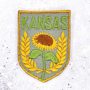 May include: Embroidered patch featuring the word "KANSAS" in green, above a sunflower and wheat stalks. The patch has a gray background with a yellow border, and is shield-shaped.