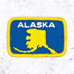 May include: A rectangular embroidered patch featuring a blue background with a yellow map of Alaska in the center. The word ALASKA is stitched in white capital letters above the map. The patch has a thick yellow stitched border.