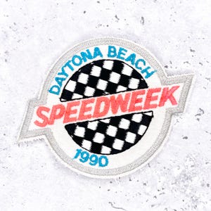 May include: Embroidered patch with a white border featuring the words "DAYTONA BEACH" in blue and "SPEEDWEEK" in red. A black and white checkered flag design is in the center, with the year "1990" in blue at the bottom. The patch is circular.
