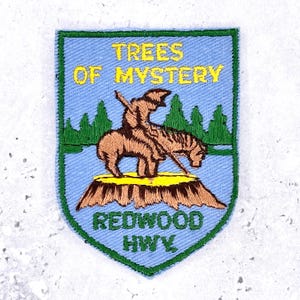 Pode incluir: Um emblema bordado em forma de escudo com fundo azul claro e borda verde. Apresenta um cavalo e cavaleiro castanhos sobre um toco bege, árvores verdes e o texto amarelo TREES OF MYSTERY acima e o texto verde REDWOOD HWY abaixo.