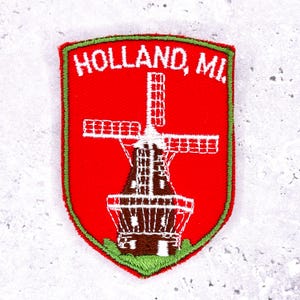 May include: Embroidered patch with a red shield shape, featuring a white windmill design and the text "HOLLAND, MI" in white. The patch has a green border.