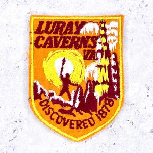 Peut inclure: Un écusson brodé en forme de bouclier sur fond orange. Il présente un texte rouge foncé indiquant LURAY CAVERNS VA et DISCOVERED 1878. Le motif inclut un soleil jaune, une silhouette de personne rouge foncé et des formations rocheuses blanches et jaunes.