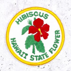 May include: A circular embroidered patch with a white background and a yellow border. The centre features a red hibiscus flower with green leaves. The green text reads HIBISCUS HAWAII STATE FLOWER, arched around the central floral design.