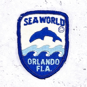 Peut inclure: Un écusson vintage SeaWorld Orlando Florida. L'écusson représente un dauphin bleu sautant au-dessus des vagues, avec les mots "SEA WORLD" et "ORLANDO FLA." en lettres blanches et bleues. L'écusson a une bordure bleue.