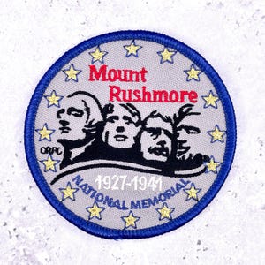 May include: A circular patch depicting Mount Rushmore National Memorial. It has a blue border, a grey background with yellow stars, and the text "Mount Rushmore" in red. The text "1927-1941 NATIONAL MEMORIAL" is in white.