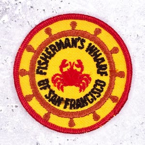 Peut inclure: Écusson circulaire avec un fond jaune, une bordure rouge et des accents et texte marron. Le texte indique "Fisherman's Wharf, San Francisco". Un crabe rouge est au centre, et une roue de navire marron entoure le texte.