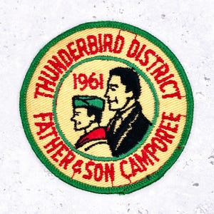 Może przedstawiać: Okrągła, haftowana naszywka z zieloną obwódką i beżowym tłem. Naszywka zawiera czerwony napis "THUNDERBIRD DISTRICT FATHER & SON CAMPOREE 1961". Przedstawia czarne i czerwone sylwetki ojca i syna.