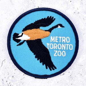 May include: Round patch with a blue background and a flying goose design. The goose has black wings, a tan body, and a white head. The text "METRO TORONTO ZOO" is in white letters.
