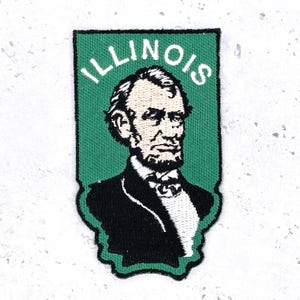 Op de afbeelding: Geborduurde patch met een portret van Abraham Lincoln, de 16e Amerikaanse president, tegen een groene achtergrond. Het woord "ILLINOIS" staat in witte letters boven het portret. De patch heeft een zwarte rand.