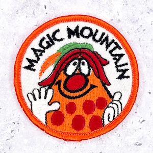 May include: A circular embroidered patch with an orange border featuring a cartoon character with an orange body, red spots, a red nose, and a green hat. The text MAGIC MOUNTAIN is embroidered in black letters along the top edge of the white background.