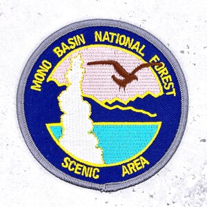 May include: Embroidered patch featuring the words "Mono Basin National Forest Scenic Area" in a circular design. The patch includes a brown bird in flight, a white rock formation, and a blue and yellow landscape.