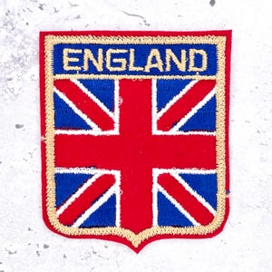 Könnte beinhalten: Ein rechteckiger gestickter Aufnäher mit der Union Jack Flagge in Rot, Weiß und Blau. Das Wort ENGLAND ist in Goldfaden auf einem blauen horizontalen Balken oben gestickt. Der gesamte Aufnäher ist mit einem dicken goldenen Rand umrandet.