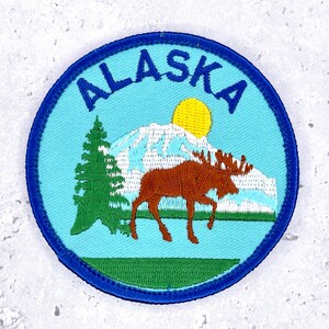 May include: A circular embroidered patch with a light blue background featuring the word ALASKA in dark blue letters at the top. The design includes a green pine tree, a brown moose, a white snow-capped mountain, and a yellow sun, all bordered in blue.