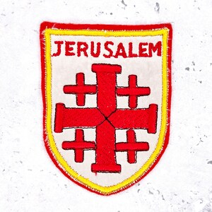 May include: Embroidered patch featuring the word "JERUSALEM" in red letters above a red Jerusalem cross on a white background. The patch has a yellow border and a red stitched edge.