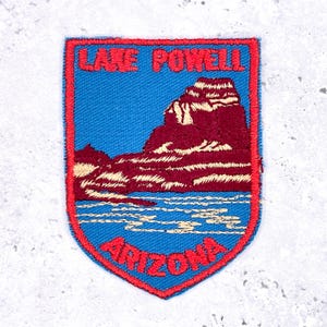 May include: Embroidered patch featuring a scenic view of Lake Powell in Arizona. The patch has a blue background with a maroon mountain and water design. The text "LAKE POWELL" and "ARIZONA" are in red.