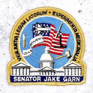 May include: Embroidered patch with a blue and gold border. The patch features an American flag, a building, and the text "SENATOR JAKE GARN". Additional text includes Latin phrases.