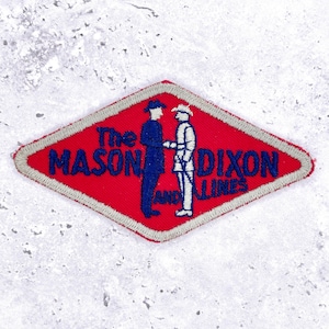 Op de afbeelding: Een rood, ruitvormig embleem met de tekst "The MASON DIXON AND LINES" in blauw. Twee silhouetten van figuren die elkaar de hand schudden. Het embleem heeft een grijze rand en is waarschijnlijk een embleem of insigne.