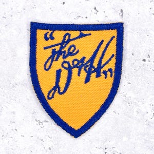 Peut inclure: Un écusson en forme de bouclier avec un centre jaune et une bordure bleue. L'écusson porte l'inscription "the Oath" en script bleu. L'écusson est probablement un emblème ou un insigne.