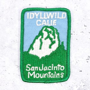 May include: A rectangular patch with a green border features the text "IDYLLWILD CALIF" above a white mountain graphic. Below the mountain is the text "San Jacinto Mountains" in white. The background is light blue.