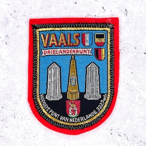 May include: A rectangular patch with a red border and a blue center. The patch features the text "VAALS" and "DRIELANDENPUNT" in gold lettering. It also includes illustrations of a tower, two pillars, and a coat of arms.