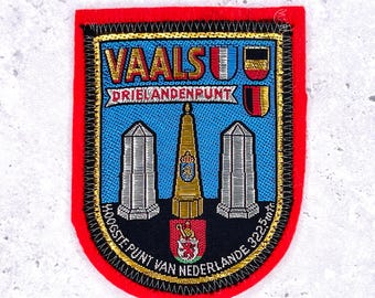 May include: A rectangular patch with a red border and a blue center. The patch features the text "VAALS" and "DRIELANDENPUNT" in gold lettering. It also includes illustrations of a tower, two pillars, and a coat of arms.