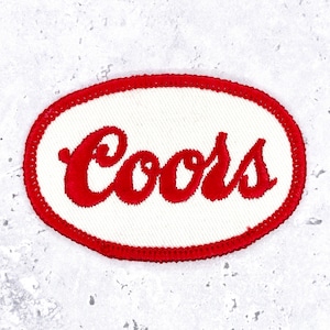 May include: An oval-shaped embroidered patch featuring a white background with a red border. The word Coors is stitched in red cursive script across the center of the patch. The design is clean and simple with a distinct red thread outline.