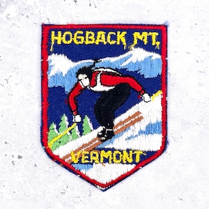 May include: A shield-shaped embroidered patch featuring a skier in a red jacket and black trousers on a snowy slope. The text HOGBACK MT. appears at the top and VERMONT at the bottom in yellow thread against a dark blue background with white mountains.