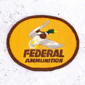 May include: An oval-shaped embroidered patch with a yellow background and a brown border. The patch features a flying mallard duck with green, brown, and white thread details. The text FEDERAL AMMUNITION is embroidered in brown below the duck.