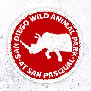 May include: A circular embroidered patch with a red background and a white border. The center features a white silhouette of a rhinoceros. The text SAN DIEGO WILD ANIMAL PARK AT SAN PASQUAL is embroidered in white capital letters around the edge.