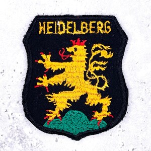 Könnte beinhalten: Ein schildförmiger, bestickter Aufnäher mit schwarzem Hintergrund. Er zeigt einen gelben Löwen mit roten Krallen und Zunge, der auf einem grünen Hügel steht. Das Wort HEIDELBERG ist in gelben Großbuchstaben oben auf den Aufnäher gestickt.