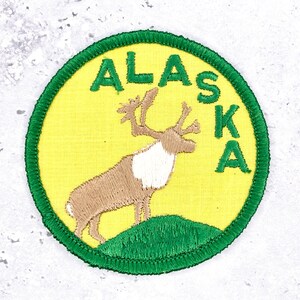 May include: A circular embroidered patch featuring a yellow background with a green border. The patch displays a brown and white caribou standing on a green hill. The word ALASKA is embroidered in green capital letters along the upper right edge.
