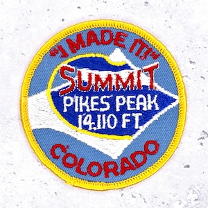 May include: Embroidered patch with a blue and white mountain design, featuring the text "SUMMIT PIKES PEAK 14,110 FT COLORADO" in red and white. The patch is circular with a yellow border and the words "I MADE IT!" in red.