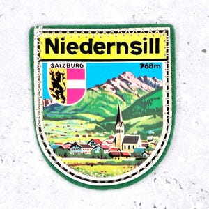 Könnte beinhalten: Ein rechteckiger Aufnäher mit dem Wort "Niedernsill" in schwarzen Buchstaben auf gelbem Hintergrund. Der Aufnäher zeigt eine farbenfrohe Illustration einer Berglandschaft, einer Kirche und den Text "Salzburg" und "768m". Der Aufnäher hat einen grünen Rand.