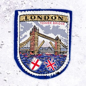 Peut inclure: Un écusson bleu représentant le Tower Bridge de Londres, avec le texte "LONDON TOWER BRIDGE". L'écusson comprend les drapeaux de l'Angleterre et du Royaume-Uni. L'écusson a une bordure blanche.