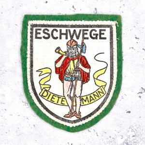 May include: A patch with a green border and a white interior. The patch features a cartoon figure in red and yellow clothing, holding a horn and a staff. The text "ESCHWEGE" and "DIETEMANN" are displayed.