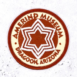 May include: A circular embroidered patch featuring a rust-colored background with a white six-pointed star design in the center. The text AMERIND MUSEUM and DRAGOON, ARIZONA is stitched in rust-colored thread along the cream-colored border.