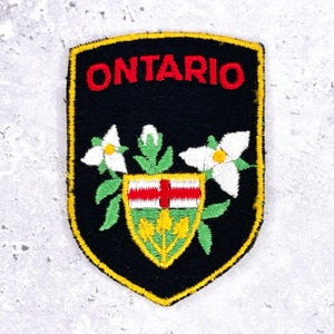 Op de afbeelding: Geborduurde patch met het woord "ONTARIO" in rood op een zwart schild. Het ontwerp bevat een wapen met een rood kruis, witte bloemen en groene bladeren, allemaal geel omlijnd.