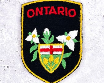 May include: Embroidered patch with the word "ONTARIO" in red on a black shield. The design includes a coat of arms with a red cross, white flowers, and green leaves, all edged in yellow.