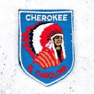 May include: A shield-shaped embroidered patch with a blue background. It features the white text CHEROKEE at the top and N CAROLINA in red at the bottom. In the center is a detailed embroidered profile of a person wearing a red and white headdress.