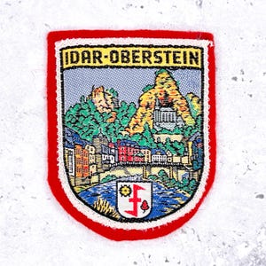 May include: Embroidered patch with a red border and a detailed landscape scene. The patch features the text "IDAR-OBERSTEIN" at the top. The scene depicts a river, buildings, a bridge, and a mountain with a castle.